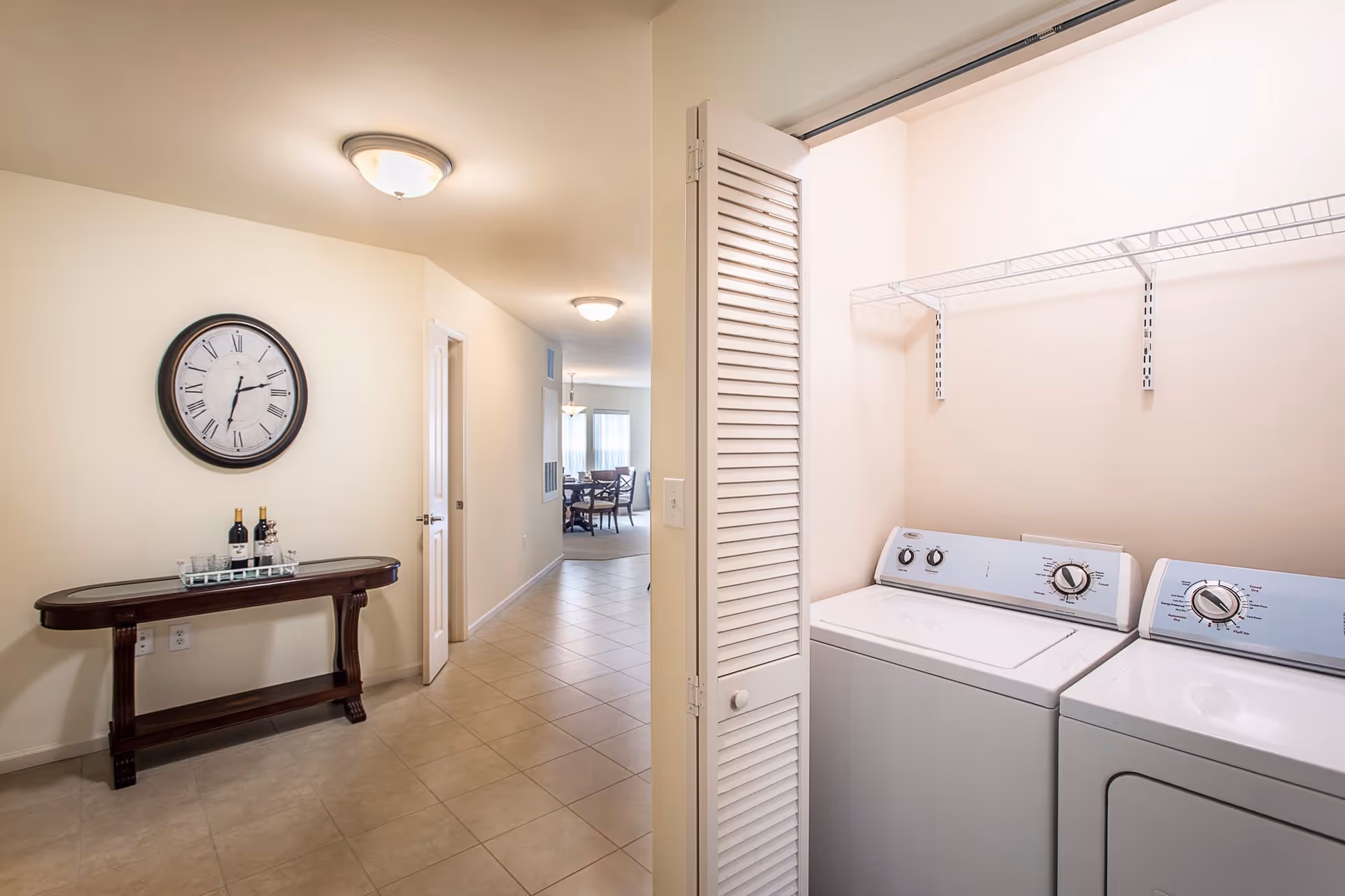 Interior view of a hallway in a senior living facility named Preston Pointe. On the right side, there is a laundry area with a washing machine and dryer behind white louvered doors. On the left side of the hallway, there is a wooden console table with two bottles of wine and glasses on a tray, and a large round wall clock above it. The hallway leads to a dining area with a table and chairs visible in the background.