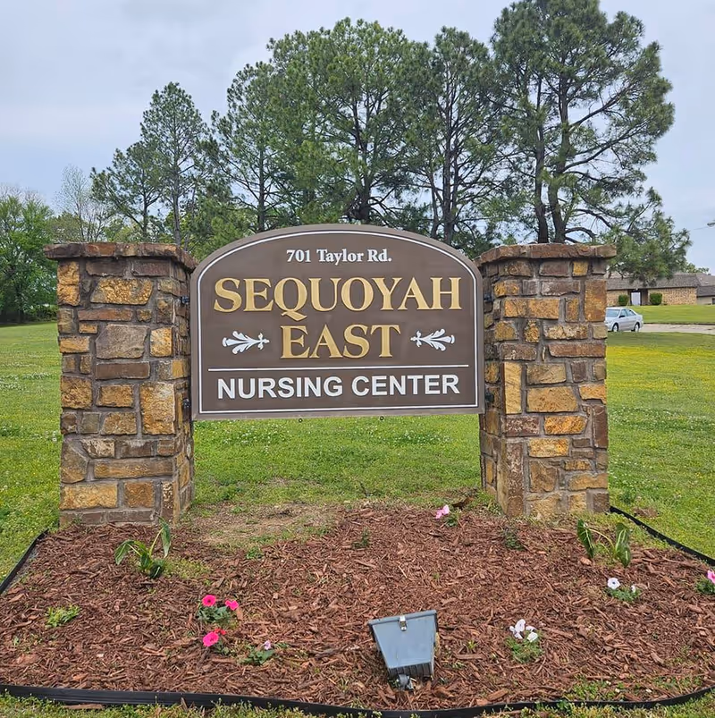 Outdoor stone sign for Sequoyah East Nursing Center at 701 Taylor Rd, surrounded by grass, flowers, and trees in the background.