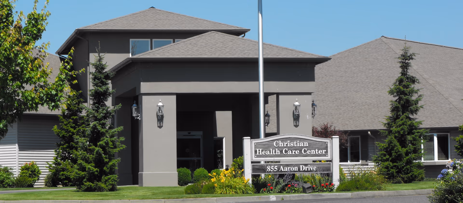 Exterior view of the Christian Health Care Center building with a sign displaying the facility name and address 855 Aaron Drive. The building has a gray facade with multiple roof peaks, surrounded by green grass, trees, and landscaping under a clear blue sky.
