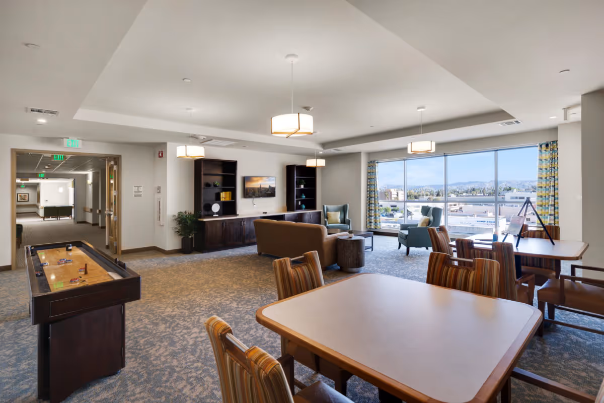 A spacious senior living common area with a shuffleboard table, several tables with striped chairs, a seating area with a brown sofa and two blue armchairs, large windows with a view of buildings and mountains, and modern ceiling lights.