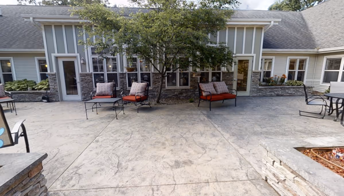 Outdoor patio area at Heritage Court with cushioned metal chairs and a small table arranged on a concrete surface. The patio is adjacent to a building with large windows, stone accents, and a tree providing some shade.