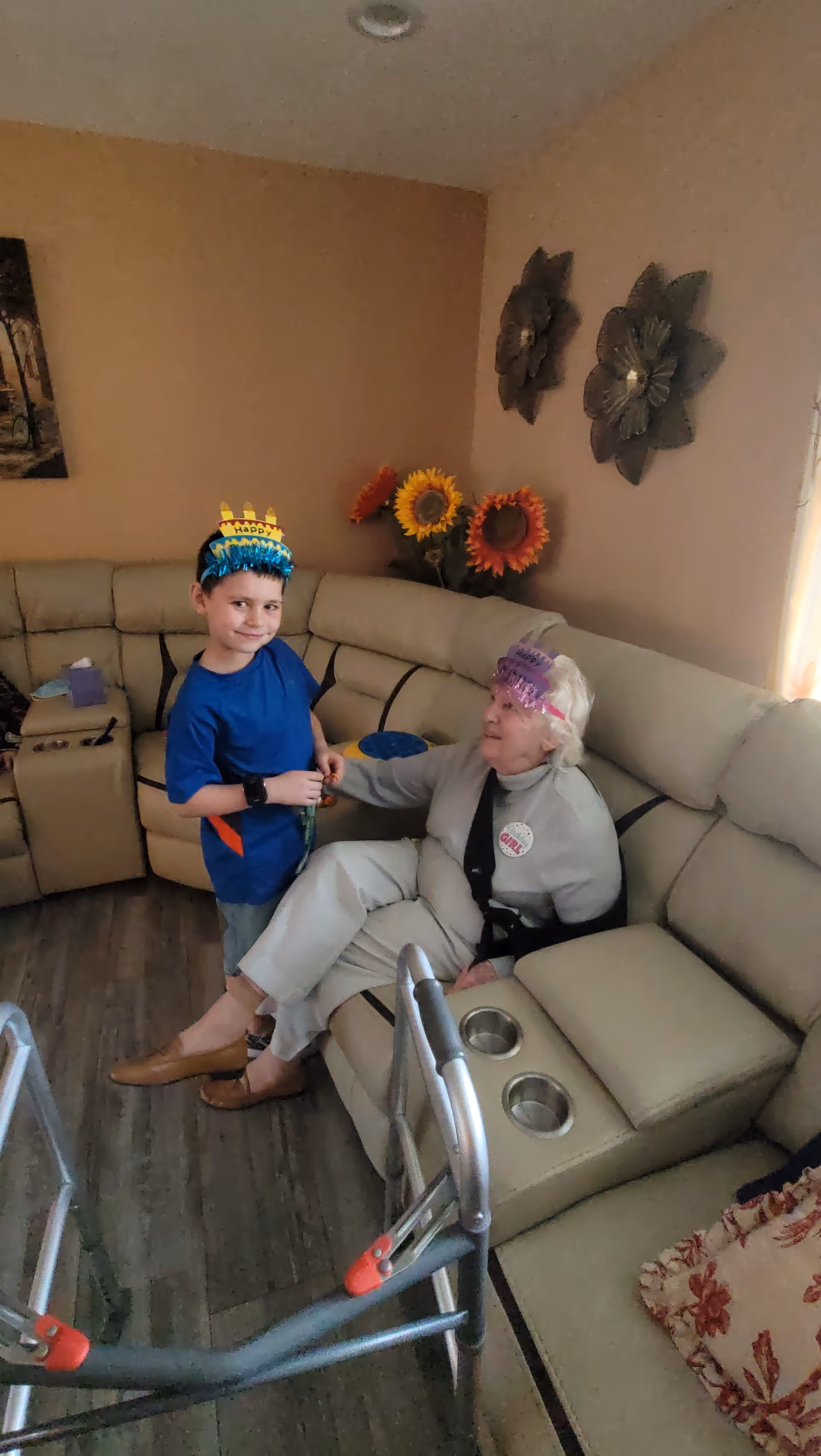 An elderly woman and a young boy wearing party crowns sit together on a beige sectional couch in a living room with sunflowers and a walker nearby.