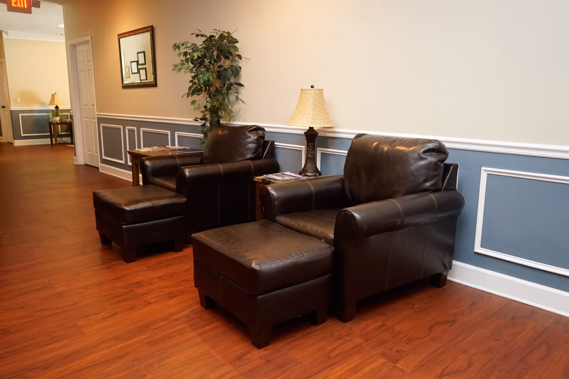 A cozy seating area in a hallway with two dark brown leather armchairs, each paired with a matching ottoman. Between the chairs is a small wooden side table with a lamp and some magazines. The walls are painted beige with blue wainscoting and white trim, and there is a framed mirror and a potted plant on the wall. The floor is a warm wood tone, and an exit sign is visible in the background.
