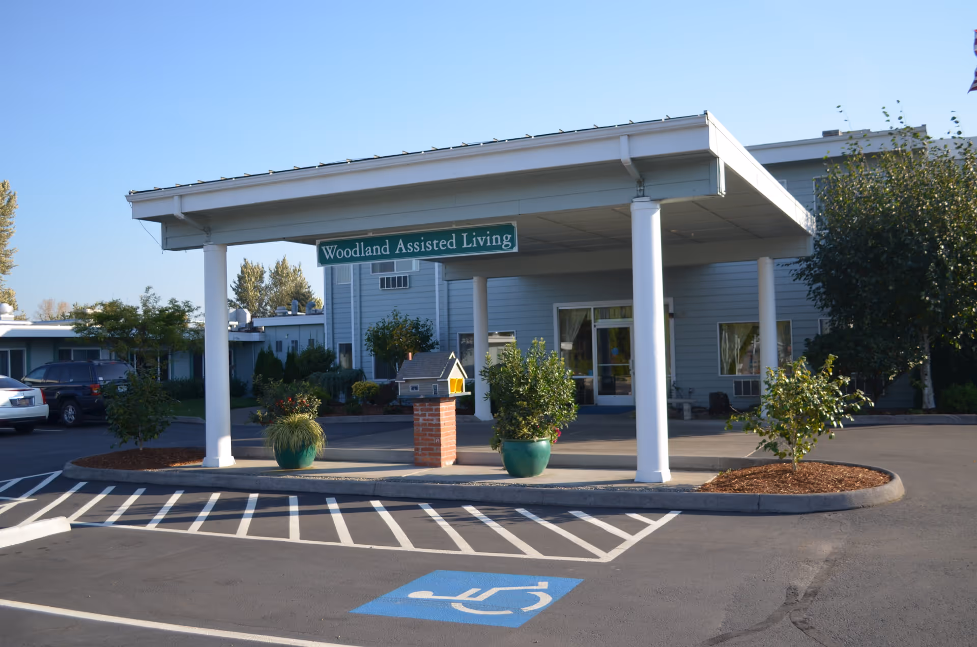 Entrance of Woodland Assisted Living facility with a covered drop-off area supported by white columns, potted plants, and a small decorative mailbox on a brick pedestal. There is a handicap parking space in front and the building is surrounded by trees and shrubs under a clear blue sky.