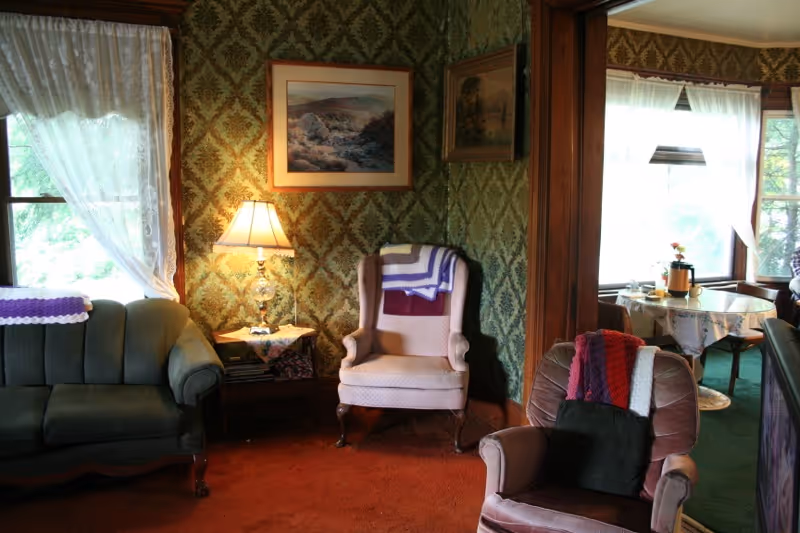 A cozy living room with vintage decor featuring a green upholstered sofa, two armchairs with crocheted blankets draped over them, a wooden side table with a lamp, and framed paintings on patterned wallpaper. Sunlight filters through lace curtains on the windows.