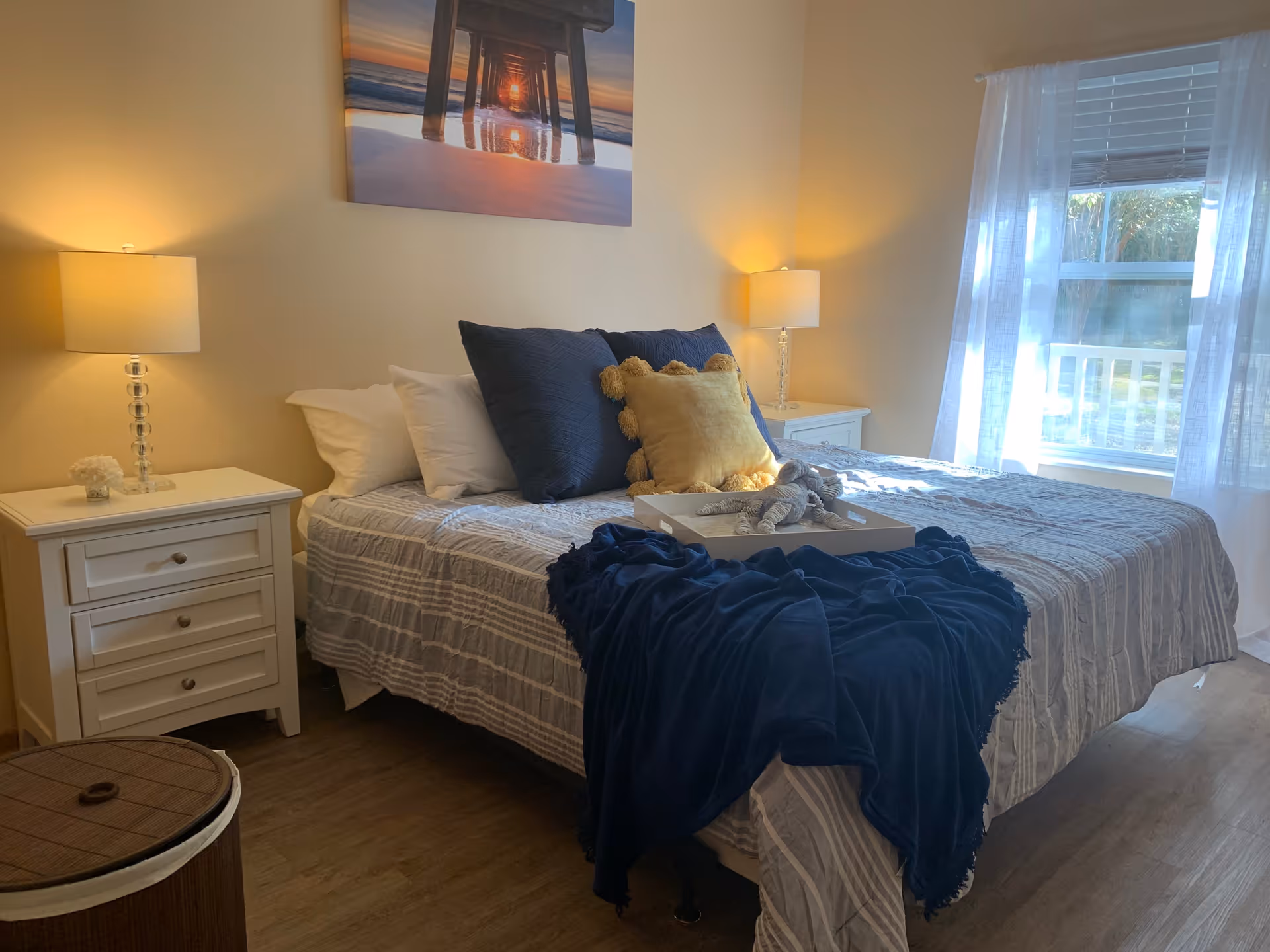 A cozy bedroom with a neatly made bed featuring white and navy blue pillows, a yellow decorative pillow, and a navy blue throw blanket. There are two white nightstands on either side of the bed, each with a lamp. A window with sheer white curtains lets in natural light, and a framed picture of a pier at sunset hangs above the bed.