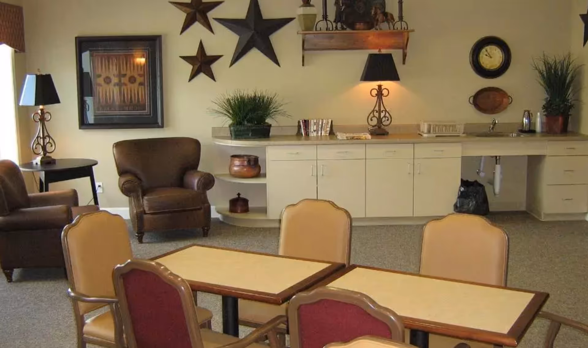 A cozy common area with a small kitchenette featuring a sink, countertop, and cabinets. There are two brown leather armchairs near a side table with a lamp. The wall is decorated with three large star-shaped ornaments, a framed picture, a clock, and a wooden shelf with decorative items. In the foreground, there are tables with beige and red cushioned chairs arranged for seating.