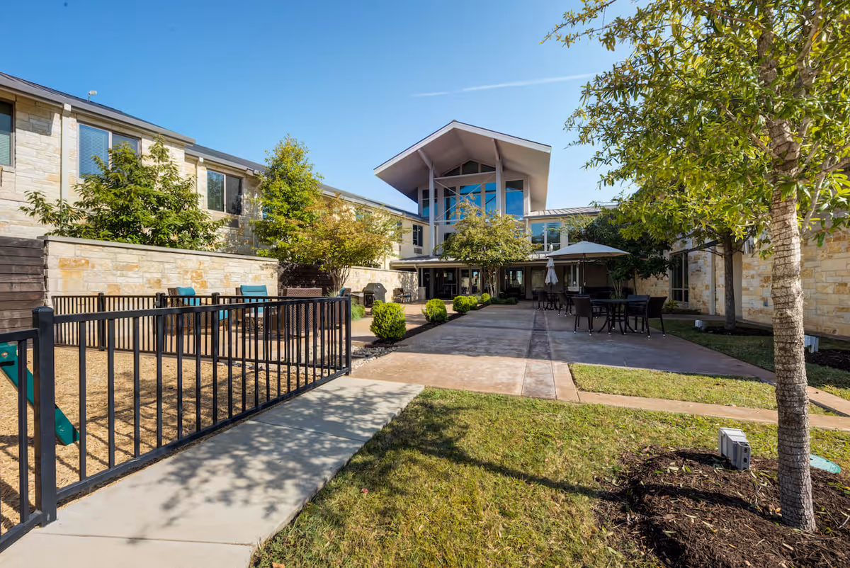 Outdoor courtyard area of Arbor Terrace Lakeway featuring a paved walkway, tables with umbrellas, chairs, green grass, trees, and a fenced-in section with benches. The building has stone walls and large windows under a clear blue sky.