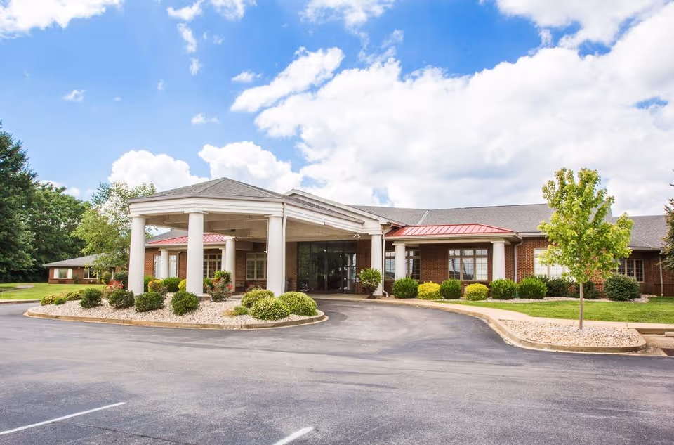 Front exterior view of Signature HealthCARE of Hart County Rehab & Wellness Center showing a single-story brick building with white columns supporting a covered entrance, surrounded by landscaped bushes and trees under a partly cloudy blue sky.