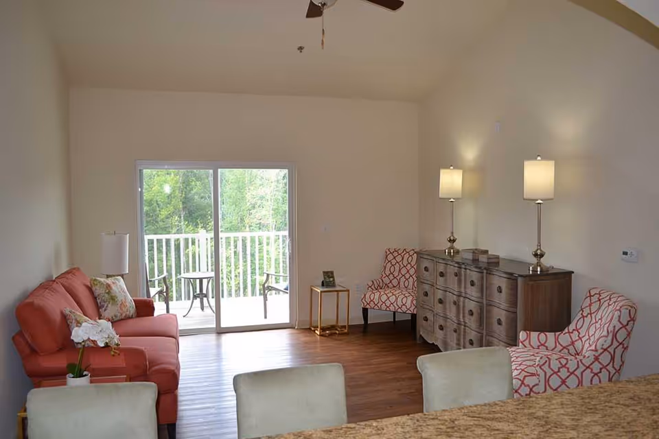 A cozy living room with a coral sofa adorned with floral pillows on the left, two patterned armchairs on the right, a wooden dresser with two lamps on top, and a small side table. Sliding glass doors lead to a balcony with outdoor chairs and a table, overlooking green trees.