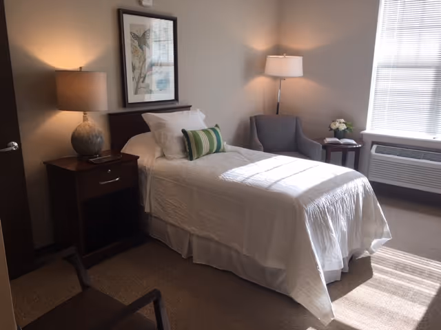 A neatly made single bed with white bedding and a green striped pillow in a well-lit bedroom. There is a wooden nightstand with a lamp and a framed butterfly picture above the bed. A gray armchair and a small round table with a flower arrangement and an open book are near a window with blinds. An air conditioning unit is below the window.