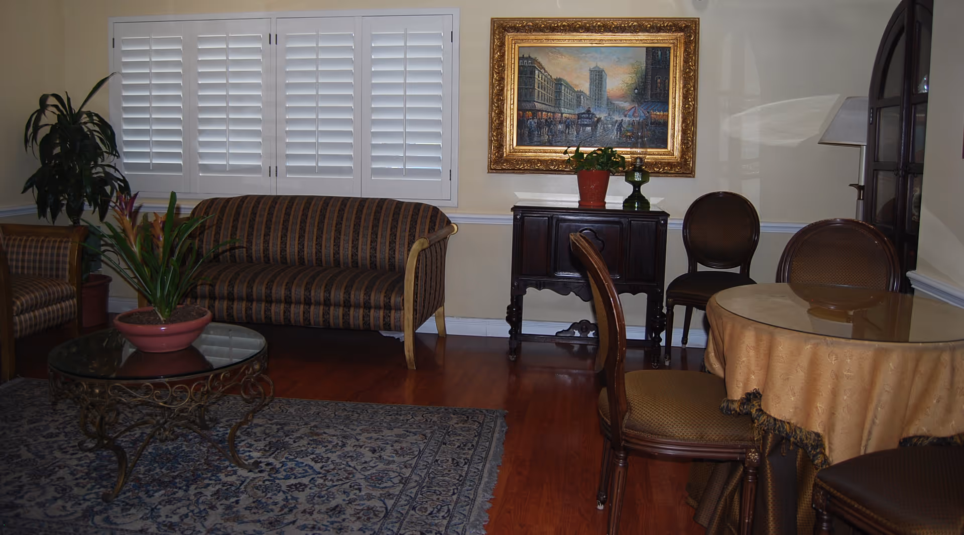 A cozy living room area with a striped sofa, a glass-top coffee table with a potted plant, a patterned rug, and a round table covered with a beige tablecloth surrounded by chairs. There is a framed painting on the wall above a small dark wooden cabinet with two plants on top. White plantation shutters cover the window, and a floor lamp is visible in the corner.