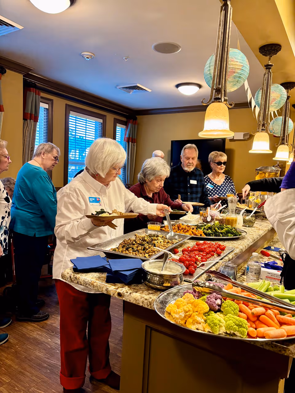 A group of elderly people serving themselves food from a buffet-style spread on a kitchen island in a well-lit room. The buffet includes various vegetables, dips, and other dishes. The room has warm yellow walls, hanging pendant lights, and windows with curtains.