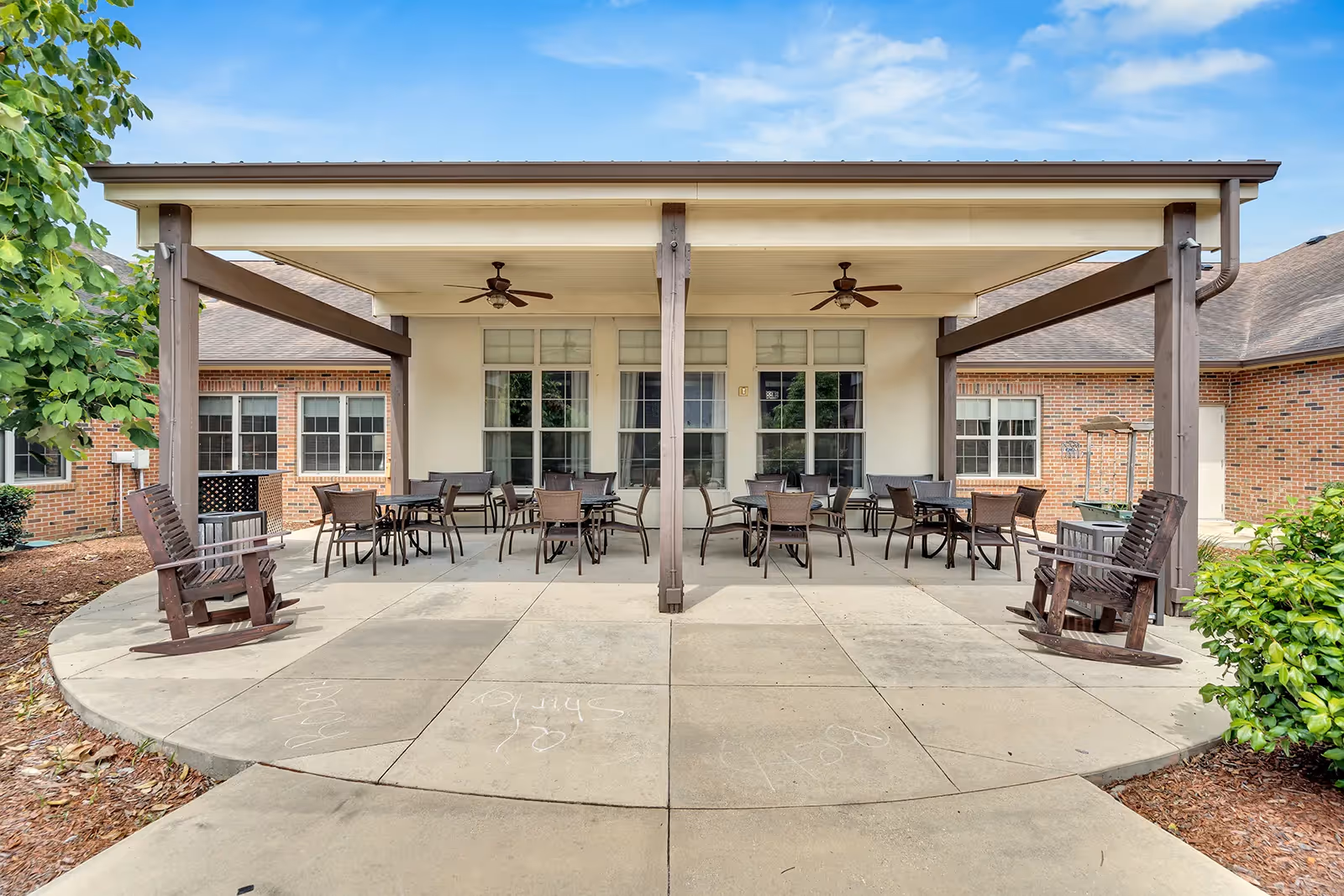 Outdoor covered patio area at a senior living facility with multiple tables and chairs arranged for seating. The patio has a concrete floor, two ceiling fans, and two wooden rocking chairs on either side. The building exterior is brick with several windows, and there are some green bushes and trees around the patio.