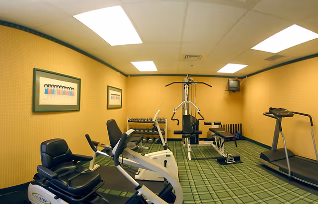 Small assisted-living fitness room with cardio machines, a weight bench, dumbbells and a TV against yellow walls and green plaid carpet.