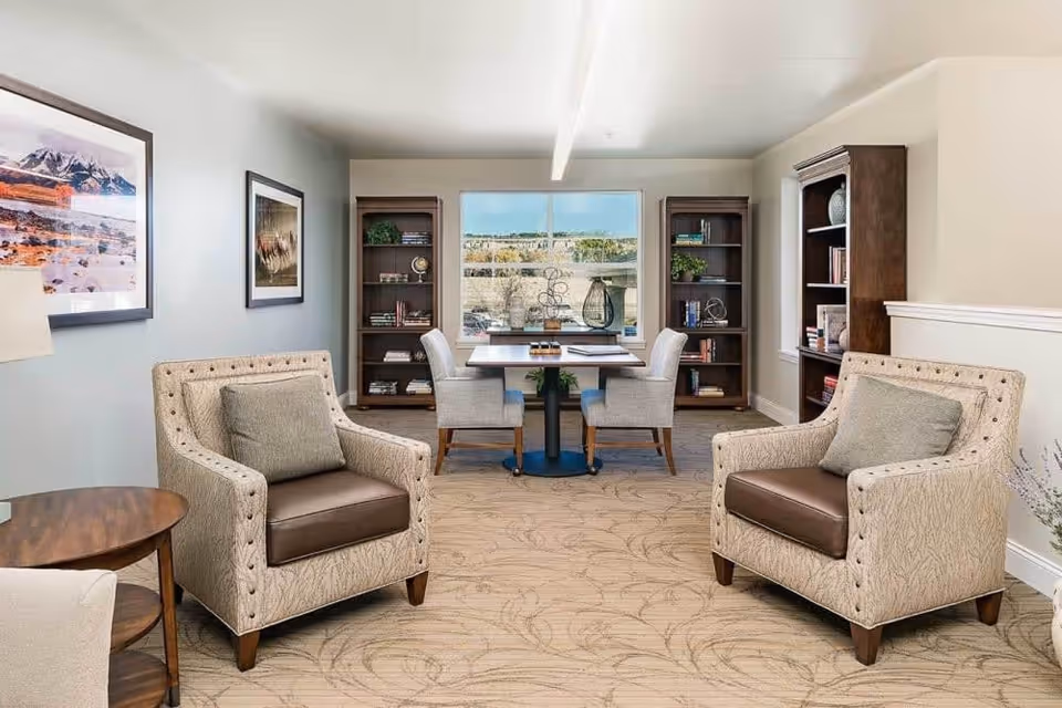 A cozy interior room with two beige armchairs featuring brown leather cushions and gray pillows facing each other. Between them is a round wooden side table with a lamp. In the background, there is a square table with four upholstered chairs, two on each side, positioned in front of a large window showing an outdoor view. Flanking the window are two tall wooden bookshelves filled with books and decorative items. The room has light-colored walls and carpet with a subtle floral pattern.