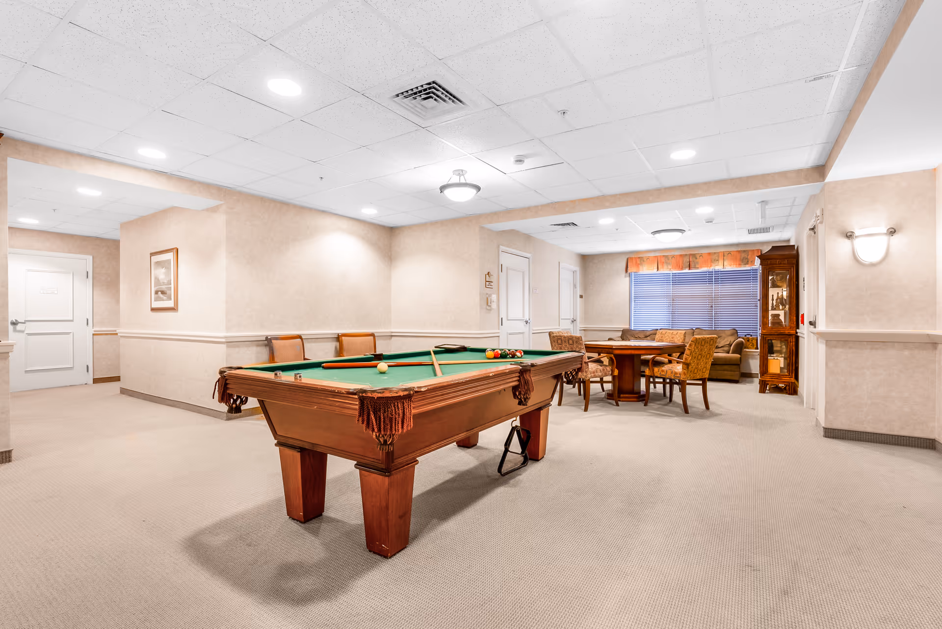 A spacious recreational room with a pool table in the center, surrounded by chairs. In the background, there is a round table with four upholstered chairs and a seating area with a sofa and a wooden cabinet. The room has beige walls, carpeted floors, and a drop ceiling with recessed lighting.