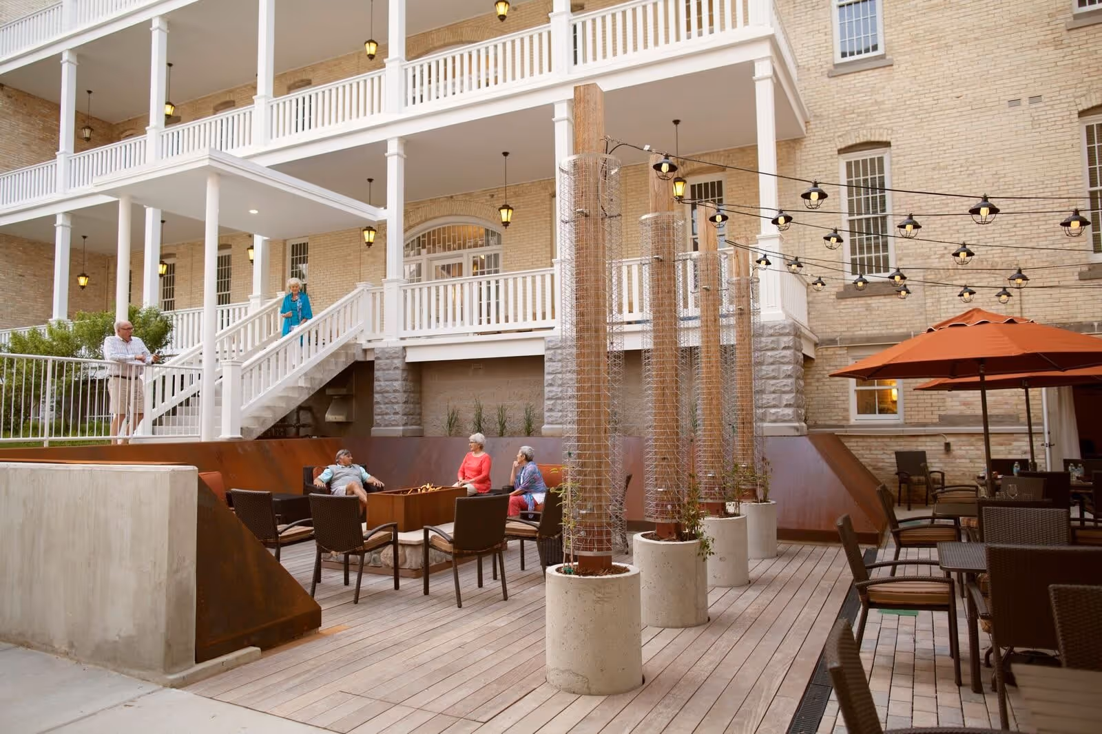 Outdoor patio area at Cordia at Grand Traverse Commons with several elderly people sitting and conversing around a fire pit. The patio features string lights overhead, potted plants, tables with chairs, and orange umbrellas. The building's brick exterior and white railings are visible in the background.