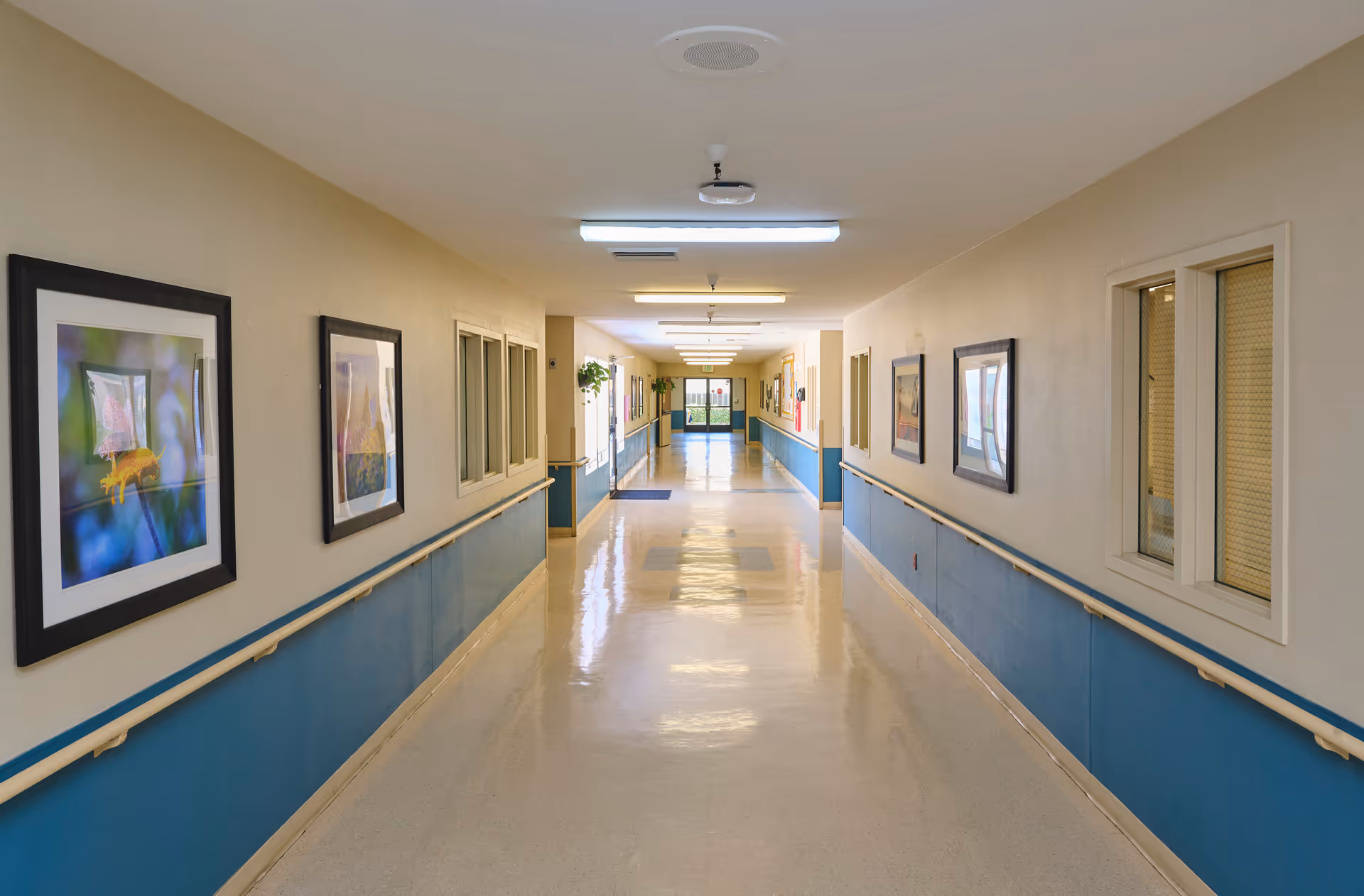 Long clean hallway in a nursing facility with handrails, framed artwork on the walls, and a polished floor leading to exit doors.