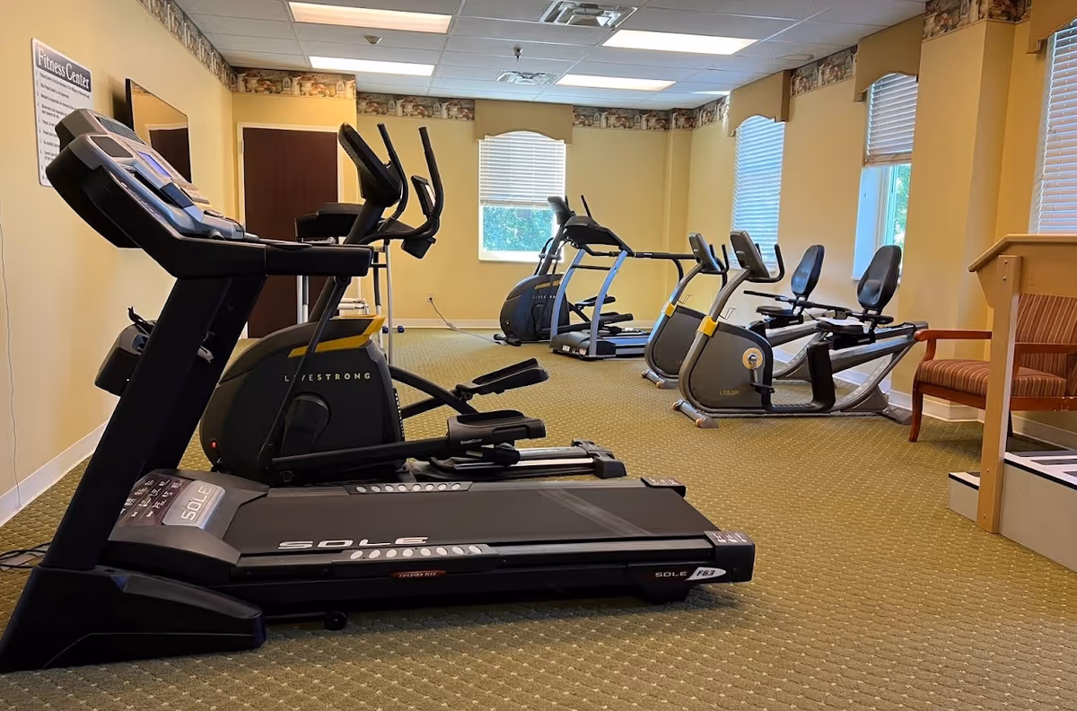 Bright fitness room with treadmills, ellipticals, and recumbent bikes in a senior living facility.