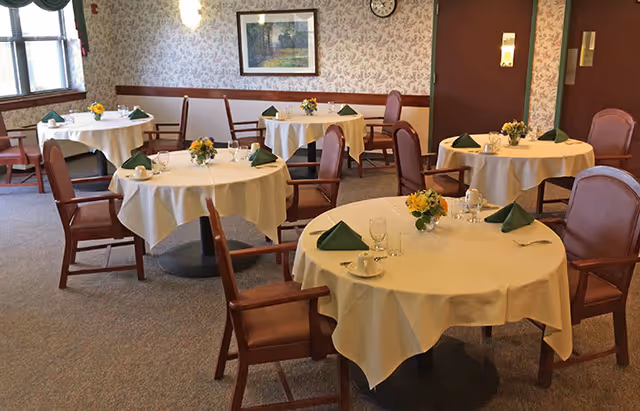 A dining room with round tables covered in white tablecloths, each set with green folded napkins, glassware, and small floral centerpieces. Wooden chairs with armrests surround the tables. The room has floral wallpaper, a framed landscape painting on the wall, a clock, and a window with green valance curtains.