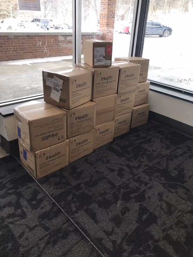 Stack of cardboard boxes labeled iHealth COVID-19 Antigen Rapid Test stacked in a corner near large windows with a view of a snowy outdoor parking area.