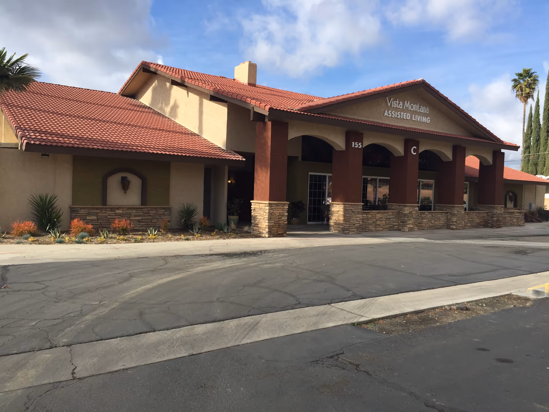 Exterior view of Vista Montana Assisted Living facility showing a building with a red tiled roof, beige and brown walls, stone accents, and a driveway in front under a partly cloudy sky.