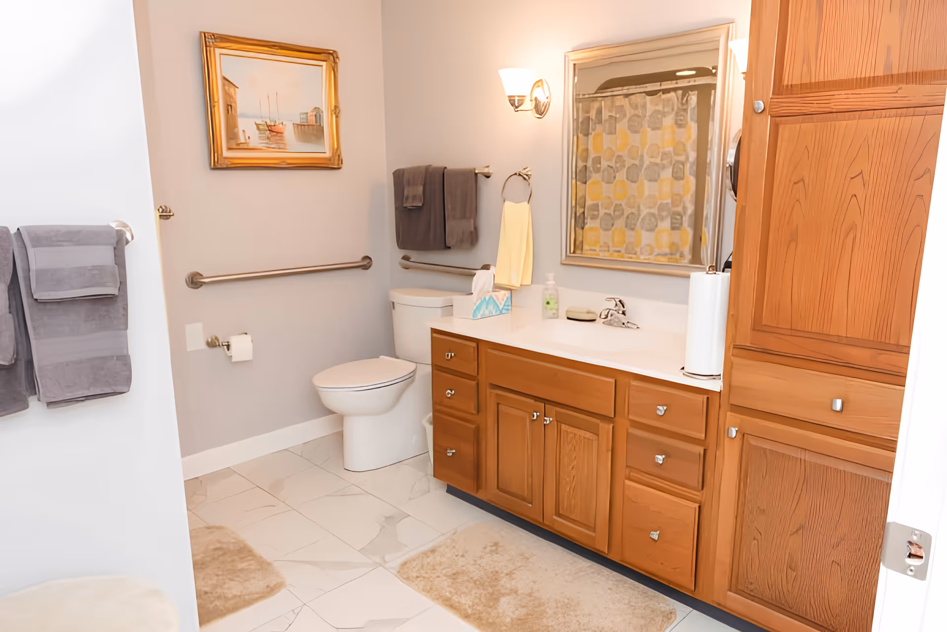 A clean bathroom with a white toilet, wooden cabinets with drawers, a countertop with a sink, a mirror above the sink, and a wall-mounted light fixture. There are gray towels hanging on a towel rack, a yellow hand towel on a ring, a framed painting on the wall, and a shower curtain with a geometric pattern visible in the mirror. The floor is tiled with light-colored tiles and there are beige bath mats on the floor.