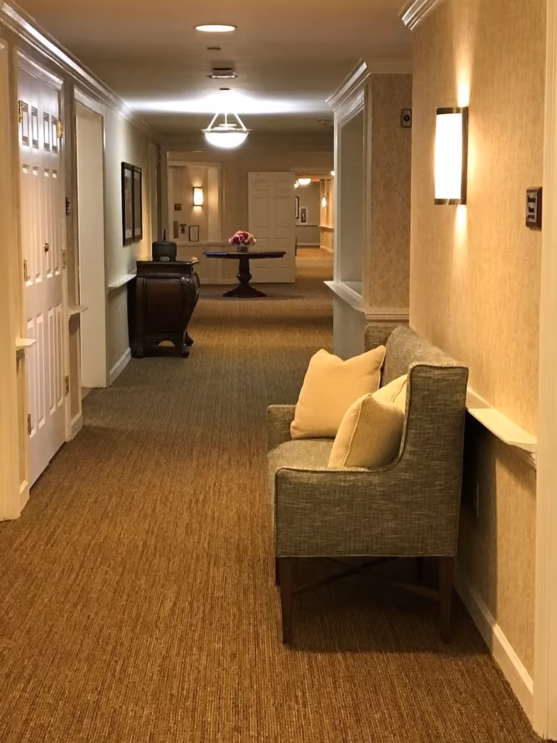 Carpeted interior hallway with a cushioned bench and pillows along the right wall and a decorative table with flowers at the far end.