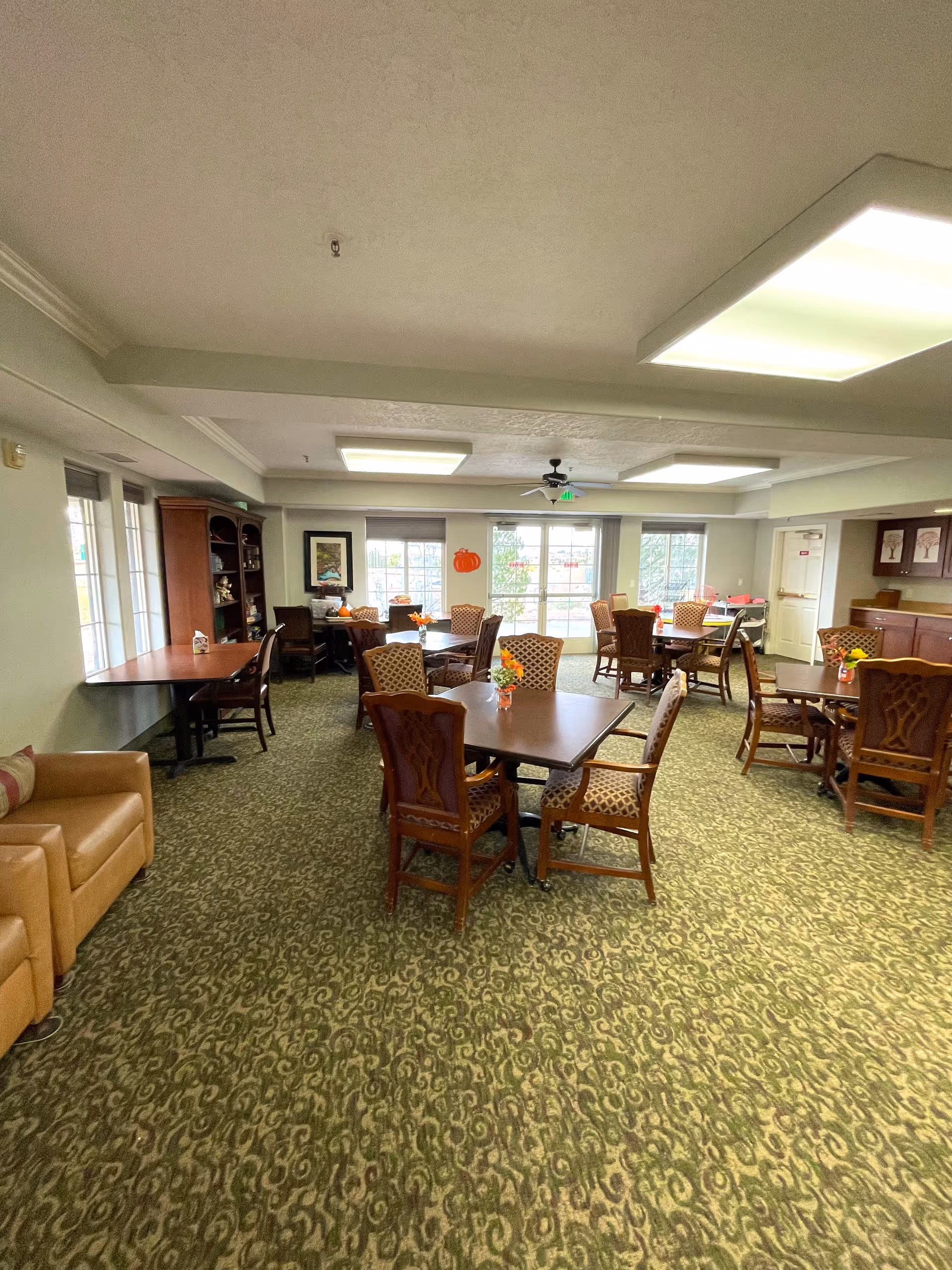 A spacious senior living dining room with multiple tables and chairs arranged neatly. The room has large windows letting in natural light, a patterned green carpet, and ceiling lights. There is a bookshelf and a tan leather armchair on the left side, and small flower arrangements on the tables. The room has a cozy and welcoming atmosphere.