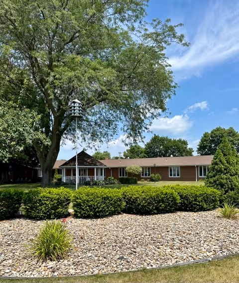 Front view of a single-story brick building with a porch, large tree and birdhouse, and landscaped shrubs and rock beds under a blue sky.