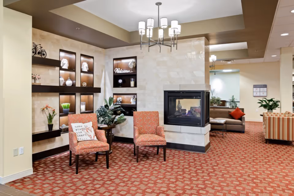 A cozy assisted living facility common area featuring two patterned orange armchairs with a small table and plants between them, a modern fireplace built into a tiled wall, decorative shelves with various ornaments, and additional seating with a brown sofa and striped armchair in the background. The space is well-lit with a chandelier and recessed lighting.