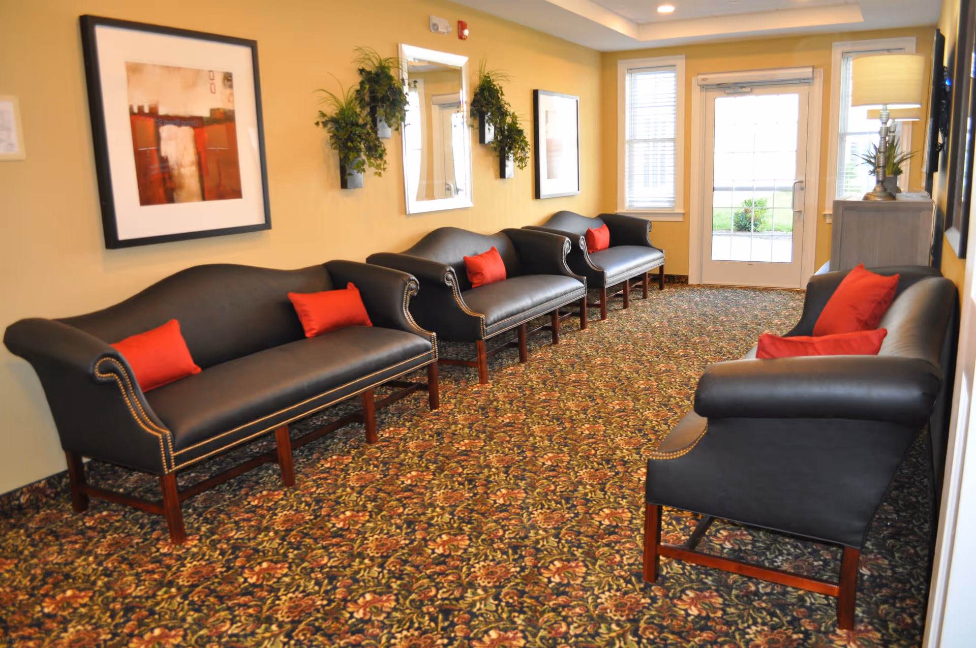 A waiting area with four black leather sofas, each adorned with red accent pillows. The room has a patterned carpet with floral designs, light yellow walls decorated with framed artwork, mirrors, and wall-mounted plants. At the far end, there is a glass door and two windows letting in natural light.