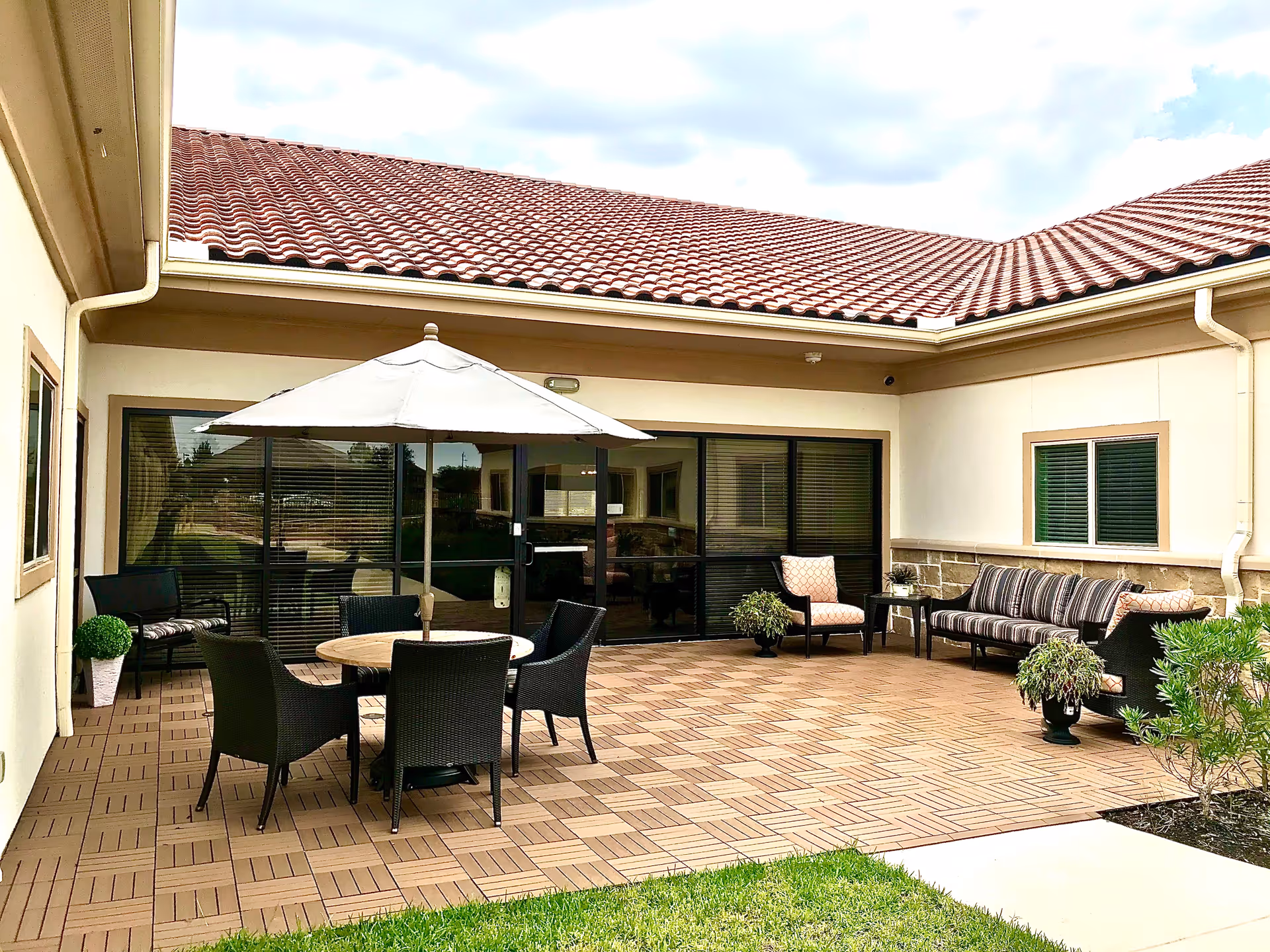Outdoor patio area with a tiled floor, a round table with an umbrella and four chairs, a cushioned sofa, two armchairs, small tables, and potted plants. The patio is surrounded by cream-colored walls with windows and a red-tiled roof under a partly cloudy sky.