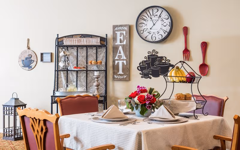 A dining table set for two with beige napkins, plates, and glasses, adorned with a floral centerpiece. Behind the table is a black metal rack holding cups and pastries, with wall decorations including a clock, a wooden sign that says 'EAT', and a wall decal of coffee cups. Two large red utensils, a fork and a spoon, hang on the wall.