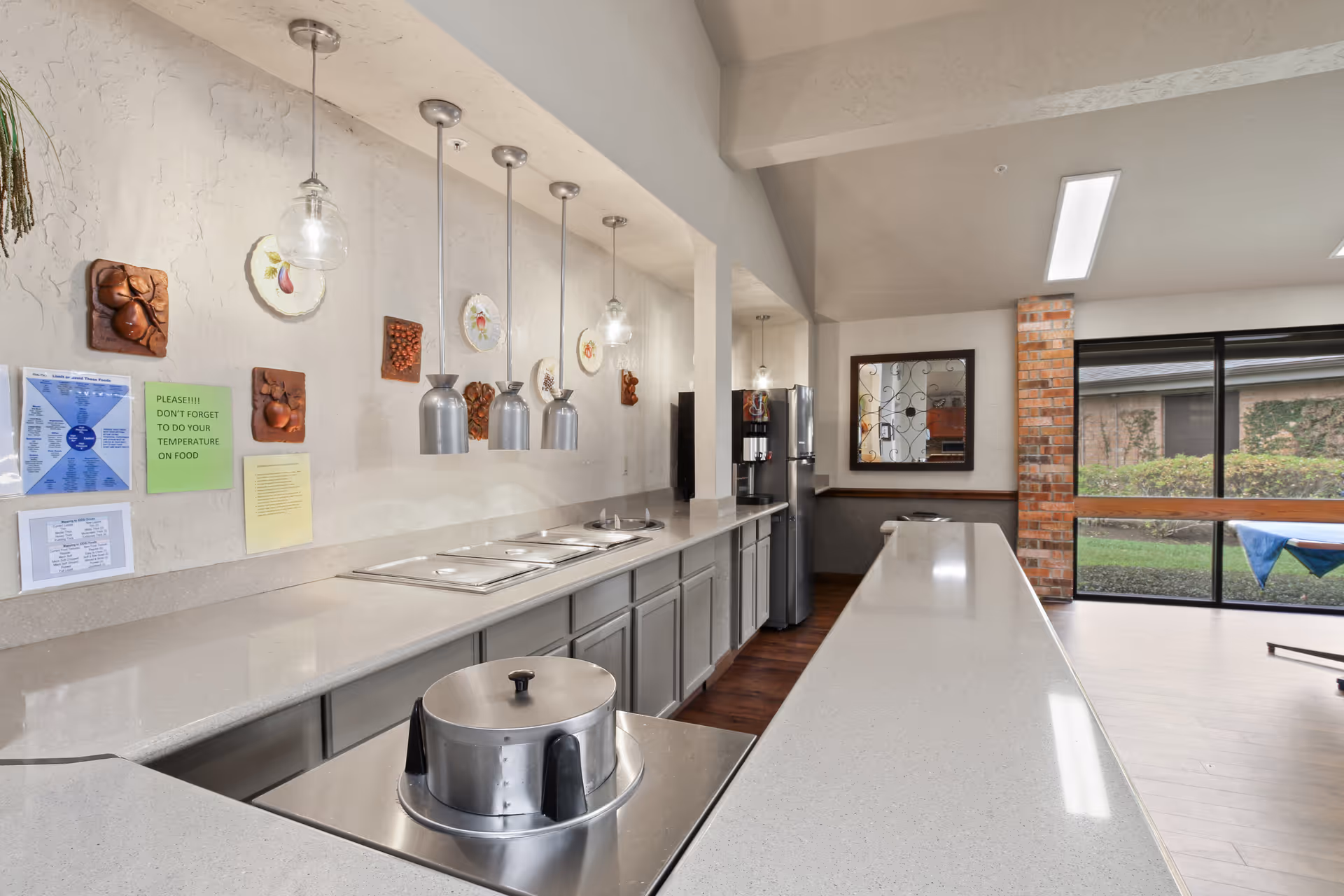 Interior view of a clean and modern kitchen area with a long countertop, stainless steel food warmers, hanging pendant lights, decorative wall plates, a refrigerator, and a large window showing an outdoor garden area.