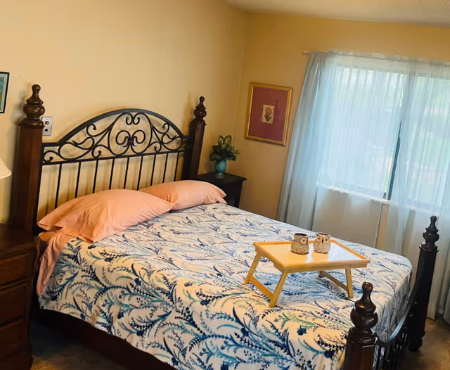 Sunlit bedroom with a decorative wooden bed, patterned blue-and-white bedspread, bedside tables, a window with sheer curtains, and a small tray on the bed.