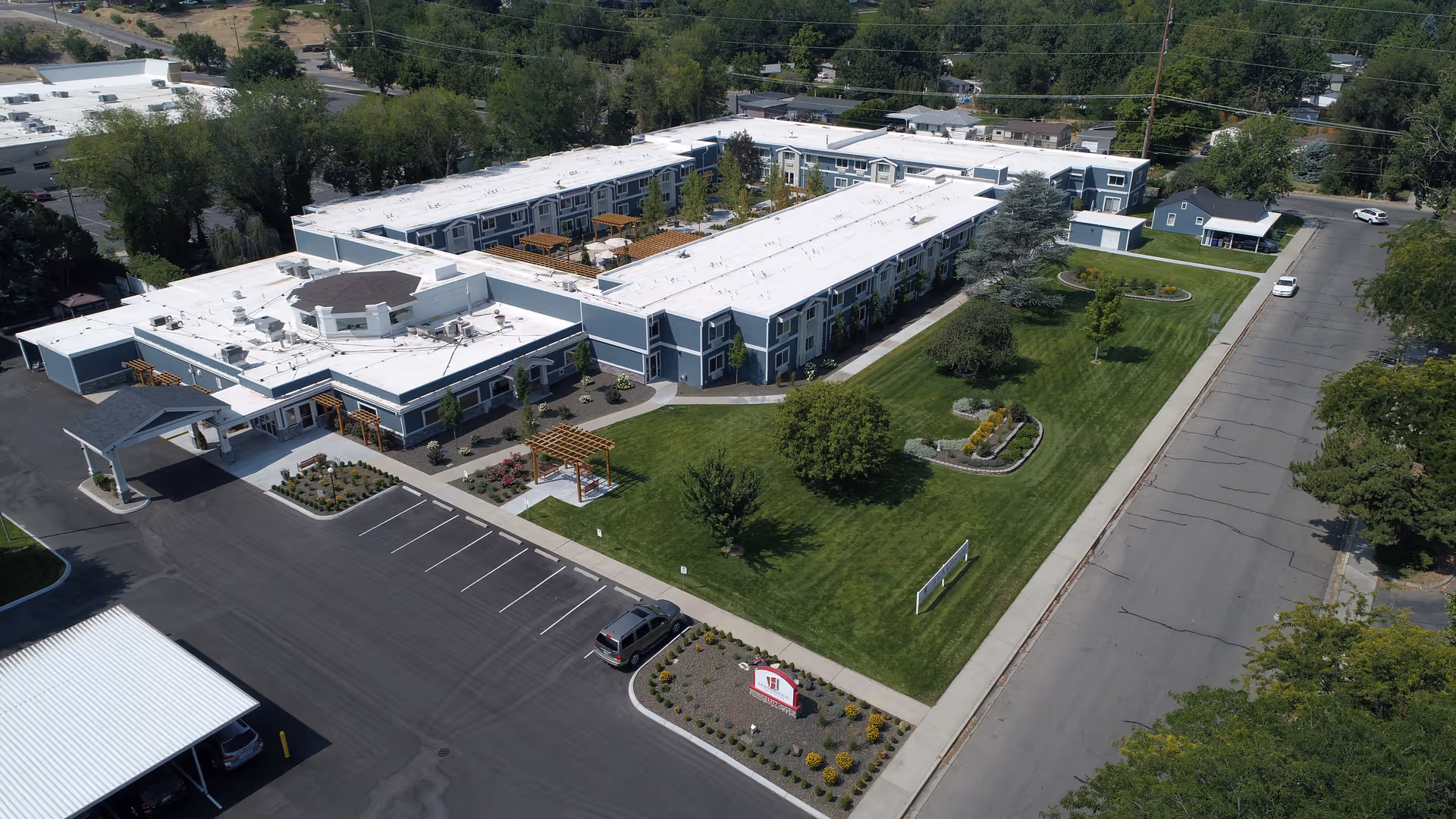 Aerial view of Heatherwood Senior Living facility showing a large, two-story building with white roofs and blue-gray exterior walls. The building surrounds a landscaped courtyard with green lawns, trees, and garden beds. There is a parking lot with a few cars, a covered entrance, and a street running alongside the property. The surrounding area includes trees and other buildings.