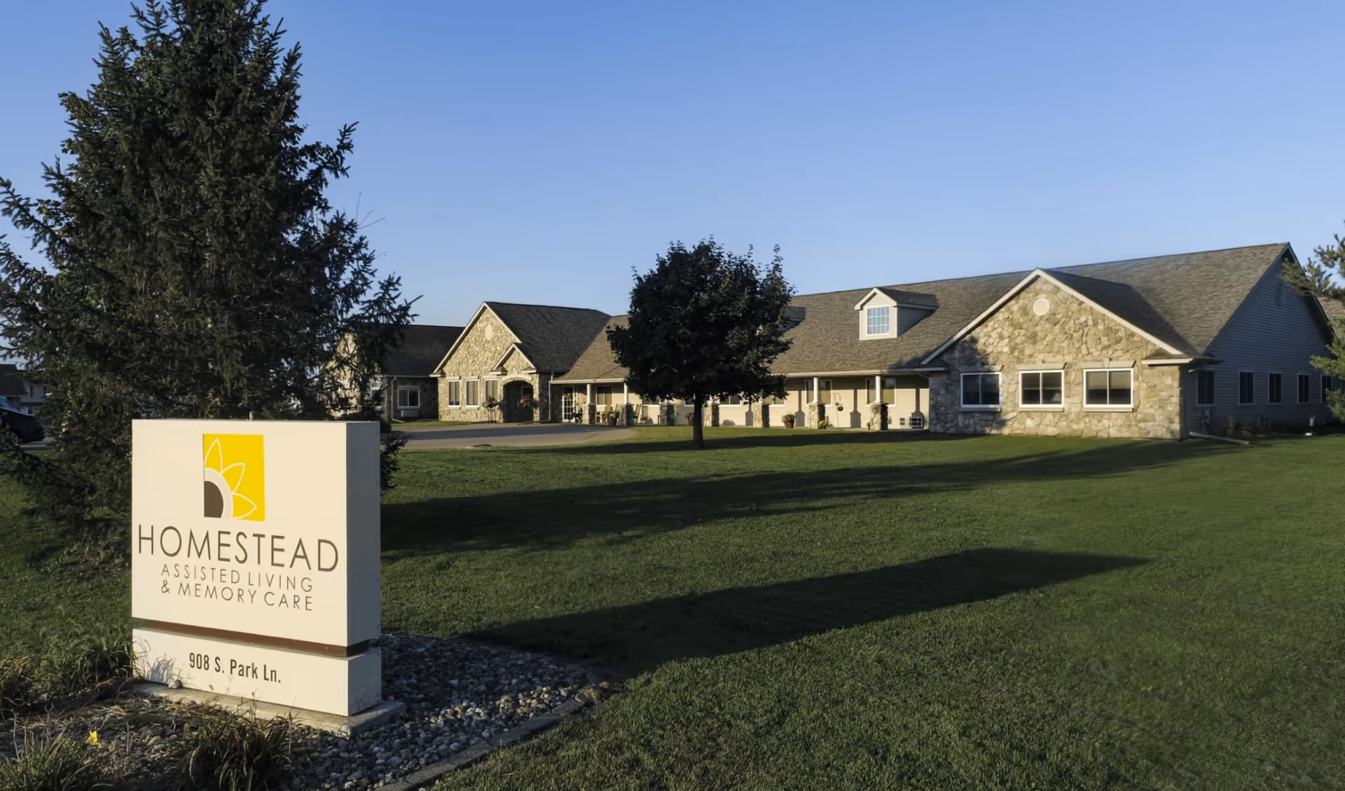 Front exterior of the Homestead Assisted Living building with a lawn and facility sign in the foreground.