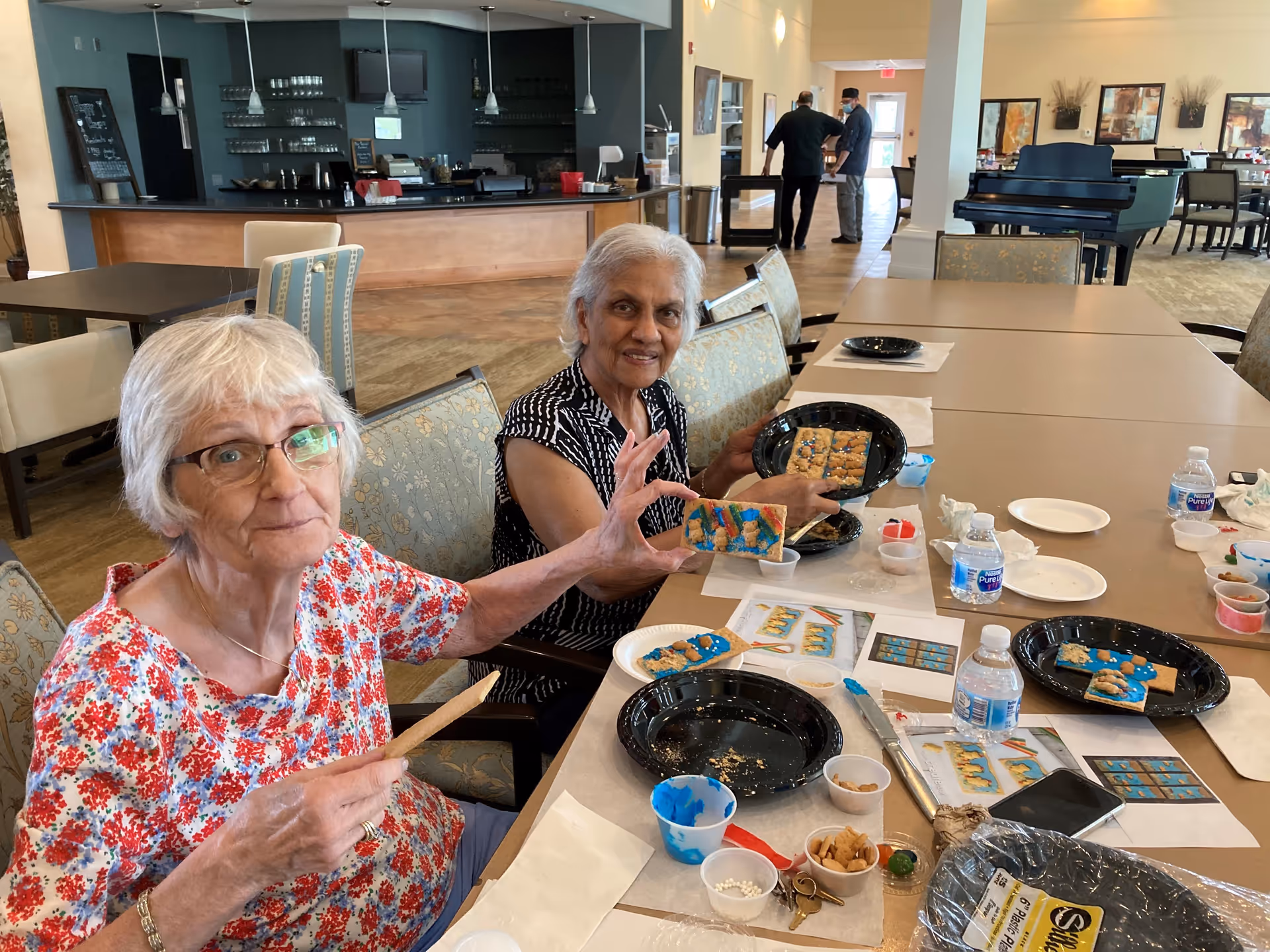 Two elderly women sitting at a table in a common area, decorating rectangular cookies with blue icing and small candies. The table is covered with plates, water bottles, and decorating supplies. In the background, there is a bar area and two men talking near the entrance, with a piano and dining tables further back.