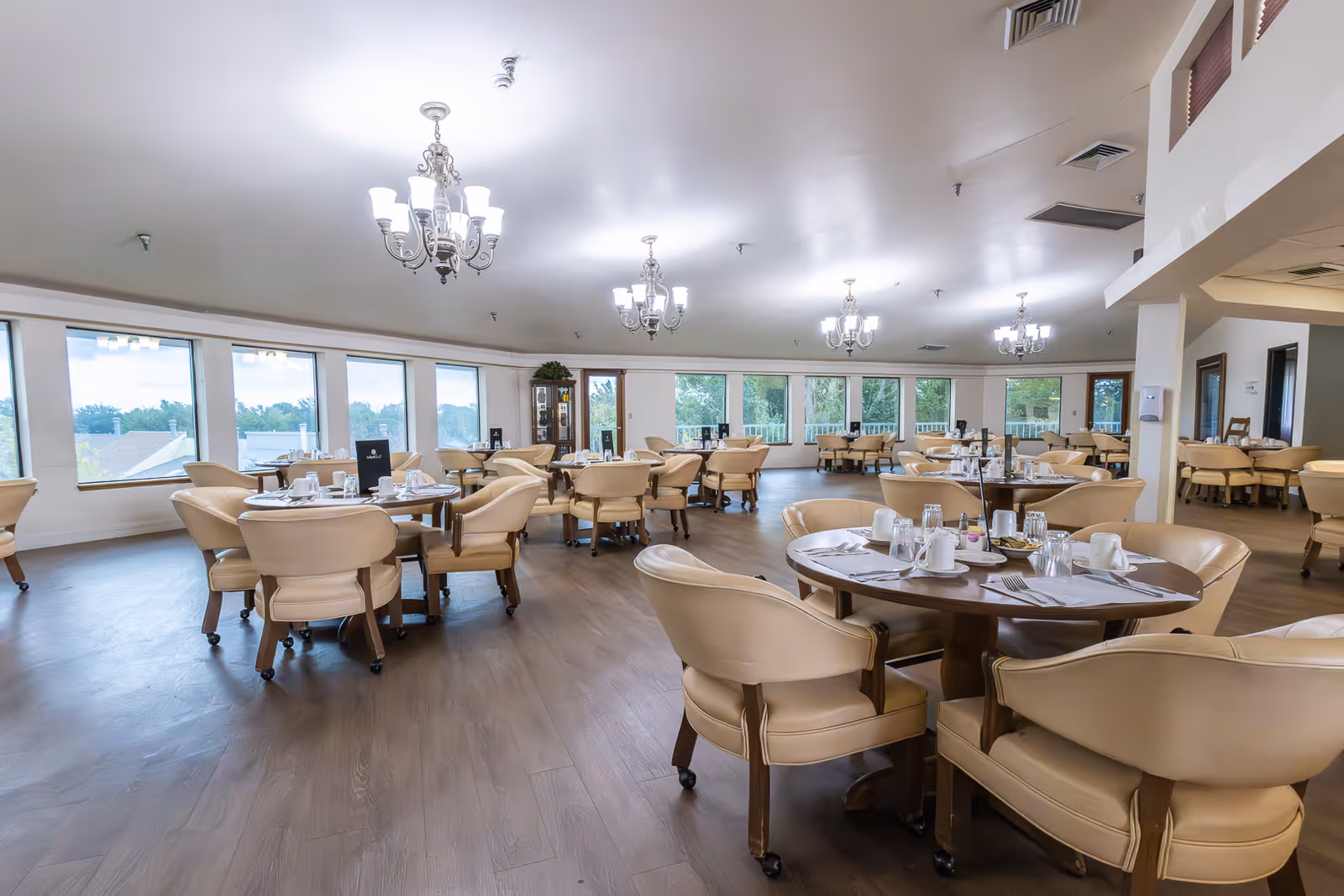A spacious dining room with multiple round tables set with white cups, glasses, and silverware. The room has large windows allowing natural light to fill the space, beige cushioned chairs around each table, wooden flooring, and several chandeliers hanging from the ceiling.