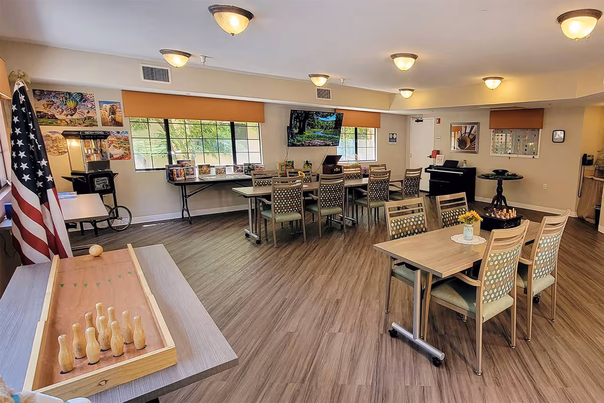 Spacious senior living common/activity room with tables and chairs, game tables, a TV, piano, and an American flag.