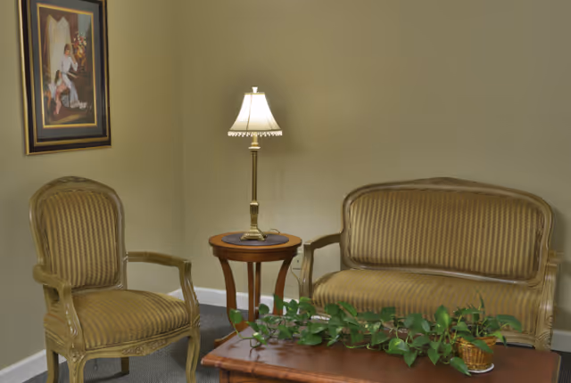 A small sitting area with gold-upholstered loveseat and armchair, a side table with a lamp, a coffee table with a potted plant, and a framed painting on the wall.