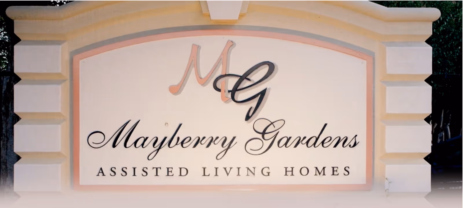 A large stone sign with the text 'Mayberry Gardens Assisted Living Homes' and stylized initials 'MG' at the top center.