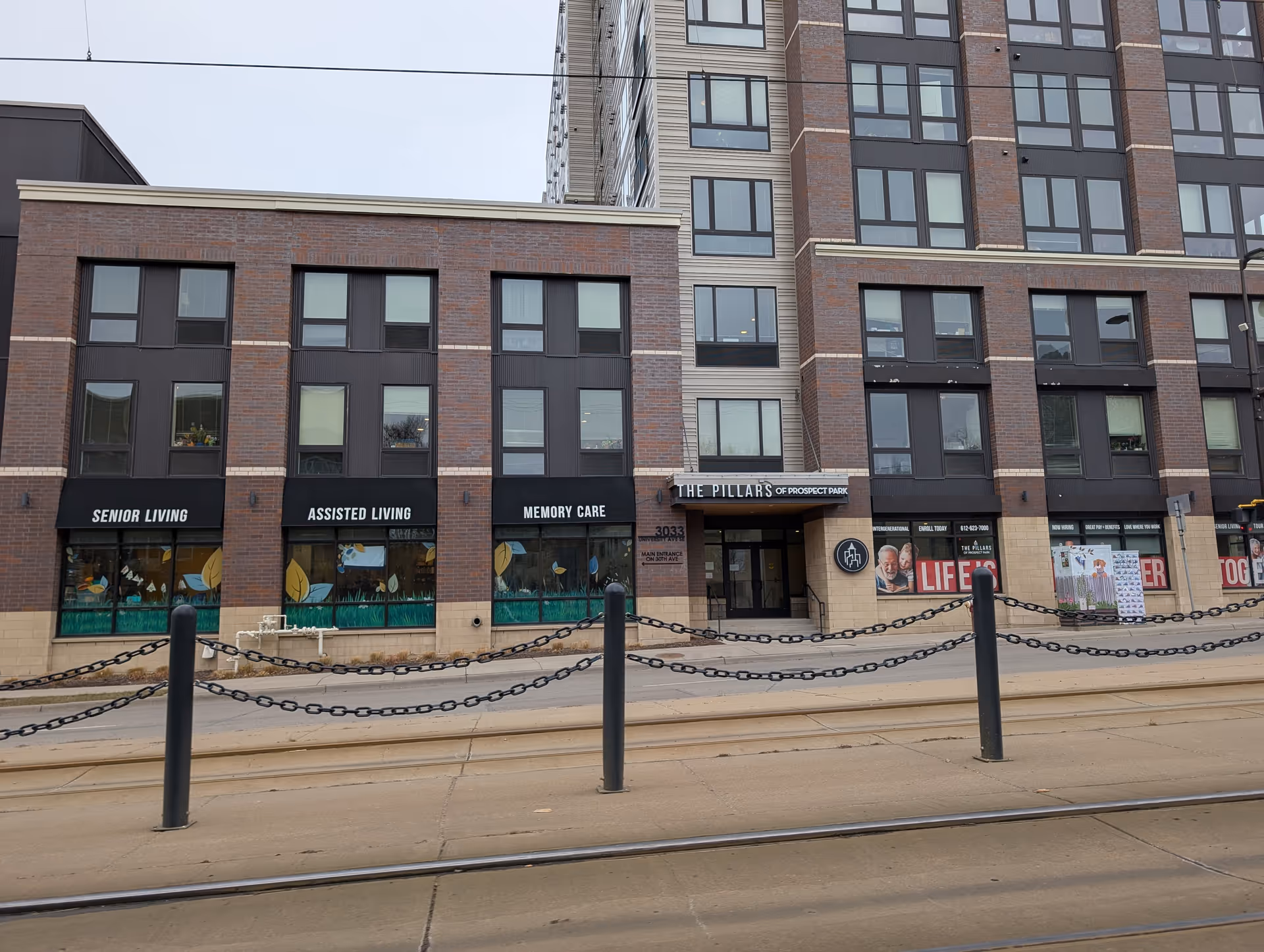 Exterior view of The Pillars of Prospect Park building showing multiple windows and entrances. The building has signs for Senior Living, Assisted Living, and Memory Care. There are black awnings above the windows and a sidewalk with black posts and chains in front.