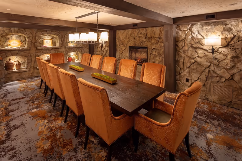 A cozy dining room with a long dark wooden table surrounded by twelve orange upholstered chairs. The room features textured stone walls with built-in shelves displaying decorative items, a fireplace, and warm lighting from a chandelier and wall sconces. The carpet has a patterned design in shades of orange and brown.