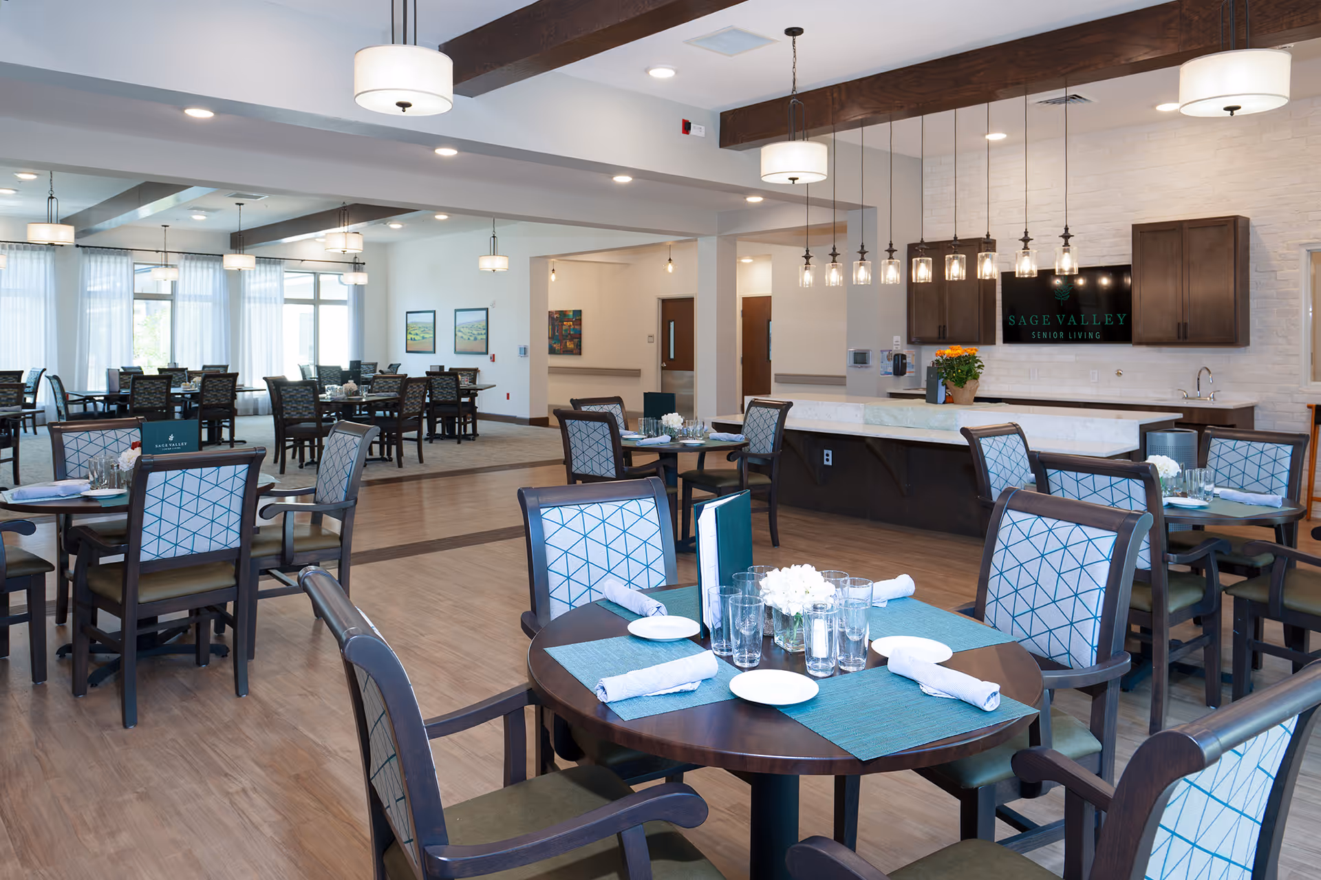 Bright, modern dining room with round tables set for meals and a serving counter displaying a Sage Valley Senior Living sign.