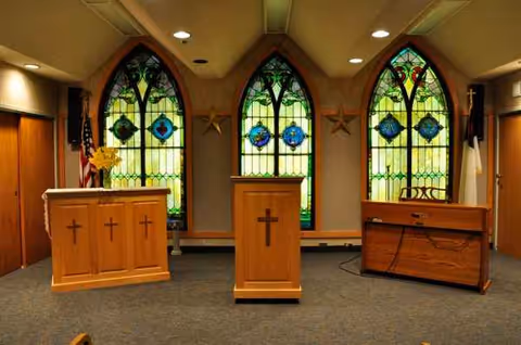 Three wooden podiums with crosses stand in a chapel-like room with stained glass windows.