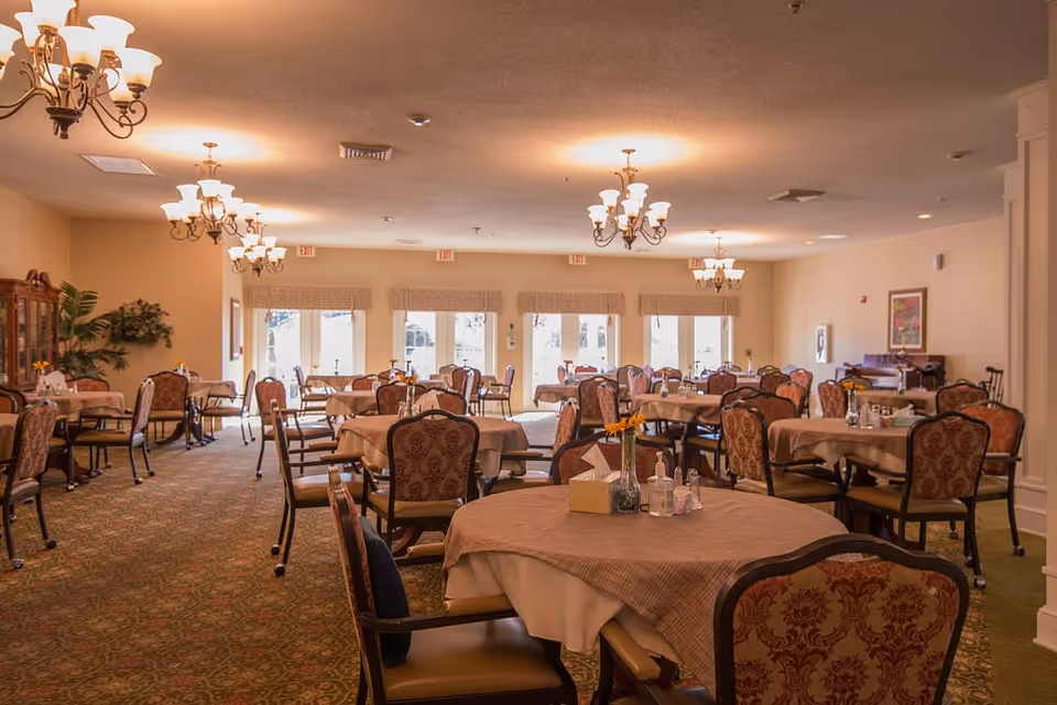 A spacious dining room in a senior living facility with multiple round tables covered with beige tablecloths. Each table is surrounded by upholstered chairs with patterned fabric. The room is well-lit with several chandeliers hanging from the ceiling. Large windows and glass doors at the far end allow natural light to enter. The carpet has a decorative pattern, and there are plants and framed artwork on the walls.