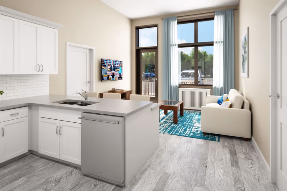 A bright and modern living space featuring a kitchen area with white cabinets, a sink, and a dishwasher. The kitchen opens into a living room with a beige sofa, a wooden coffee table on a blue patterned rug, and large windows with light blue curtains letting in natural light. A flat-screen TV is mounted on the wall opposite the sofa.